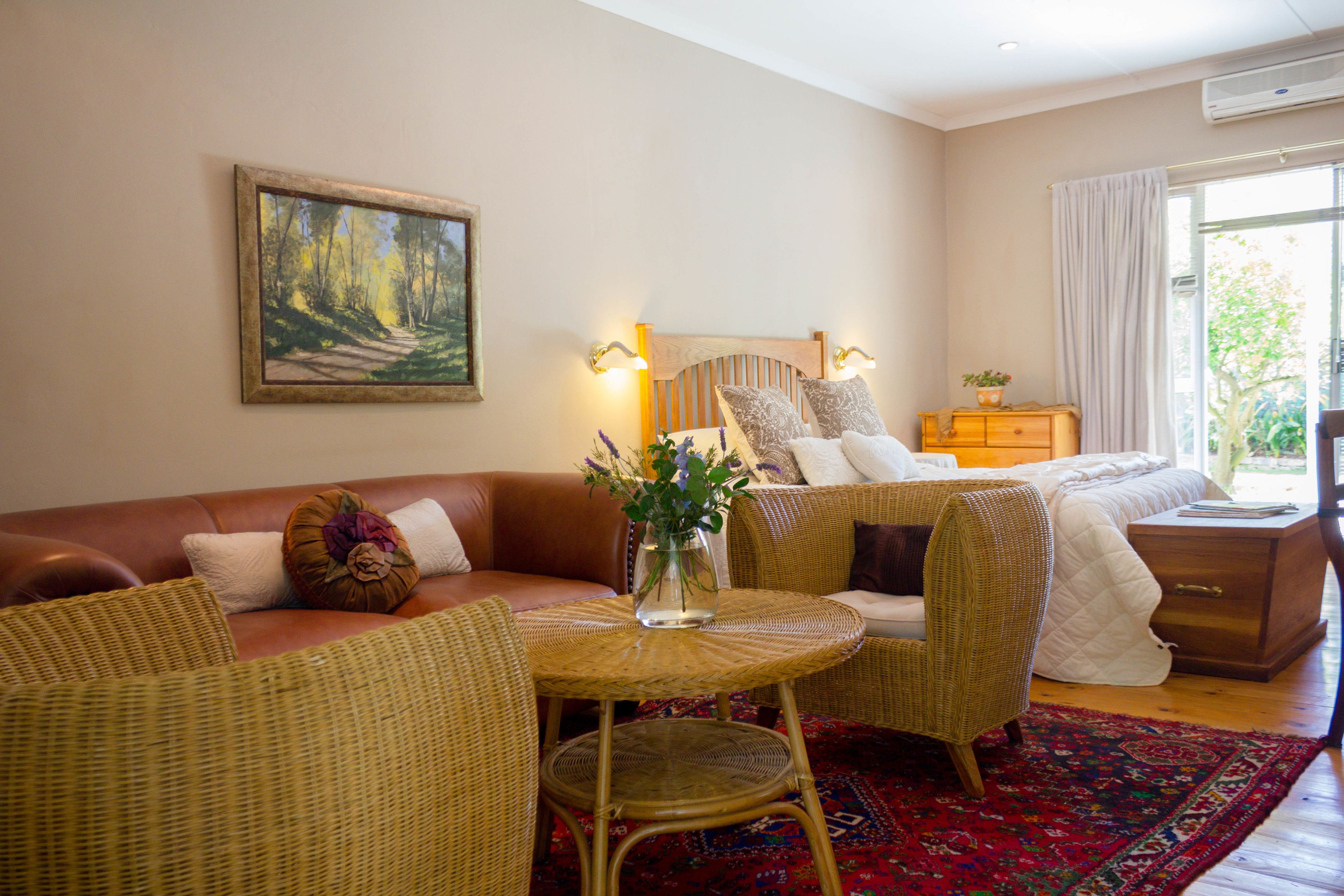 Un dormitorio con cama, zona de estar con mesa y sillas, un cuadro en la pared y una ventana con cortinas.