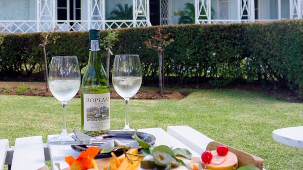 Un montaje para picnic con una botella de vino Boplaas, dos copas, y una variedad de quesos, frutas y nueces sobre una tabla de madera.