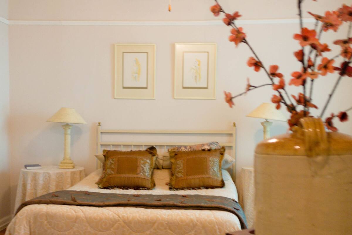 A bedroom with a bed, a ceiling fan, two framed pictures on the wall, a vase with flowers, and a patterned rug.