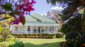 Una casa de dos pisos con techo verde, rodeada de un jardín bien cuidado con diversas plantas y césped.