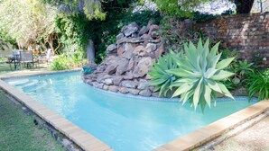 Una piscina rectangular con borde de ladrillo, rodeada por un césped y un muro de piedra.