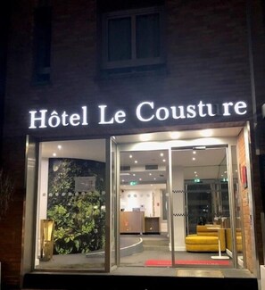 The entrance of Hôtel Le Cousture with a glass facade, a reception area visible inside, and a small outdoor seating area with yellow cushions. - Hôtel Le Cousture (Toulouse)