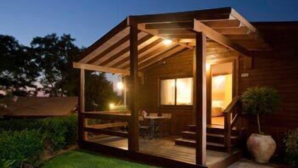 A wooden cabin with a covered porch, a staircase leading to the main entrance, and a large window. - Ramot Resort Hotel (Ramot)