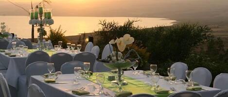 An outdoor dining area with tables set for a meal, overlooking a body of water and a mountainous landscape.
