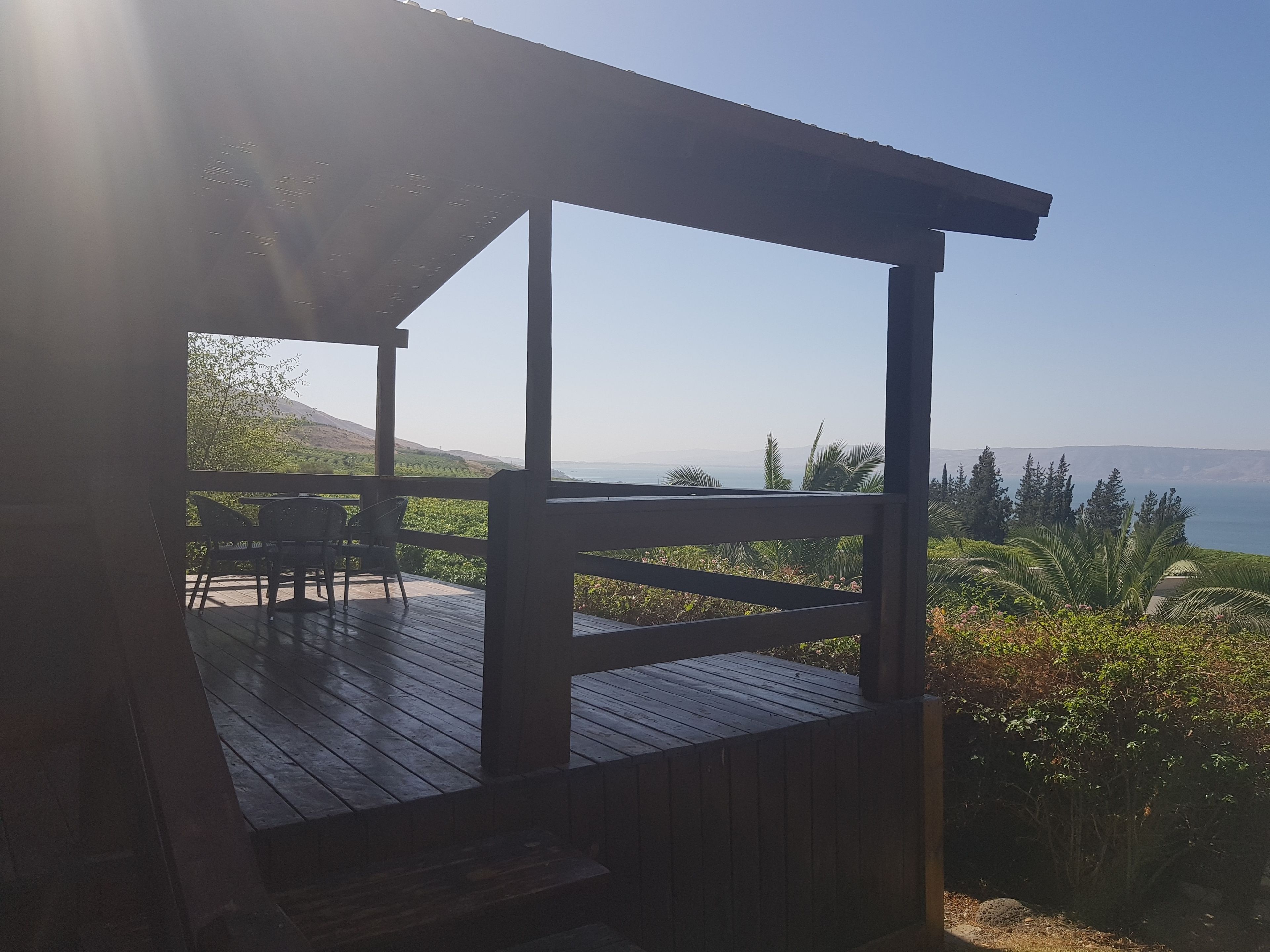 Una terraza de madera con pérgola, con vistas a un paisaje costero con vegetación y colinas lejanas.