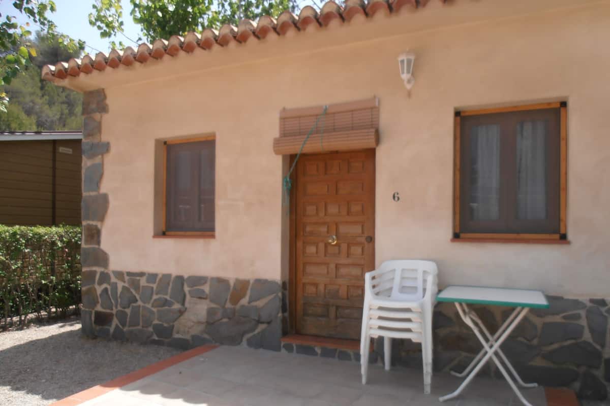 A building with a stone base and a tiled roof. There is a wooden door with a window above it, and a small table with a chair outside.