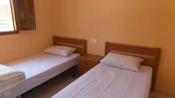 Deux lits simples avec des têtes de lit en bois, dans une pièce aux murs unis et dotée d’une fenêtre.