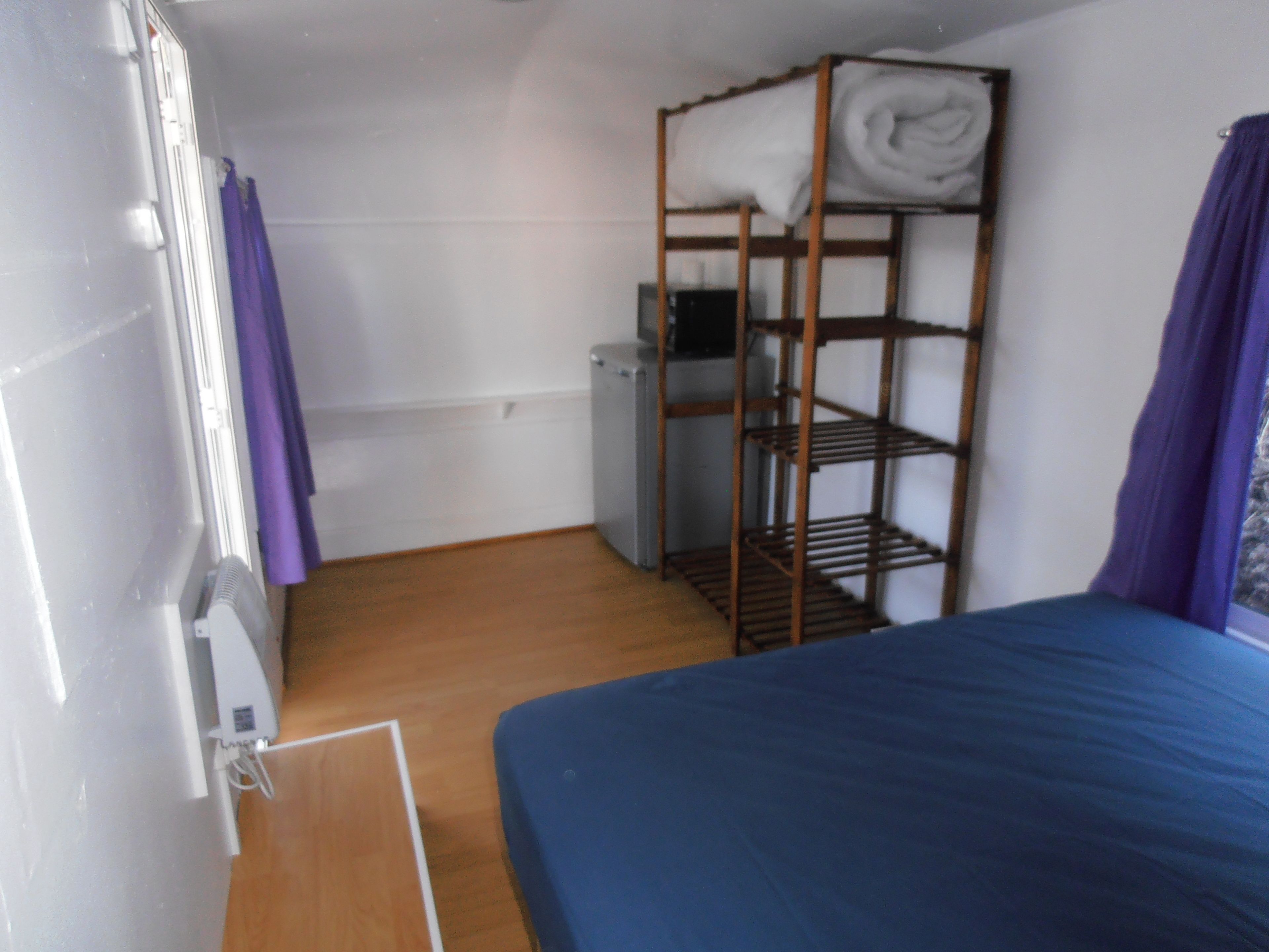 A small, compact room with a bunk bed, a wooden shelf with towels, a heater, and a window with purple curtains.