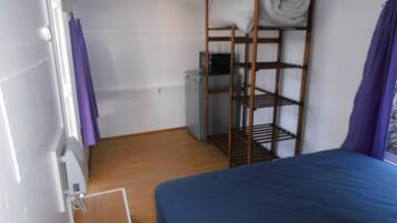 A small, compact room with a bunk bed, a wooden shelf with towels, a heater, and a window with purple curtains.