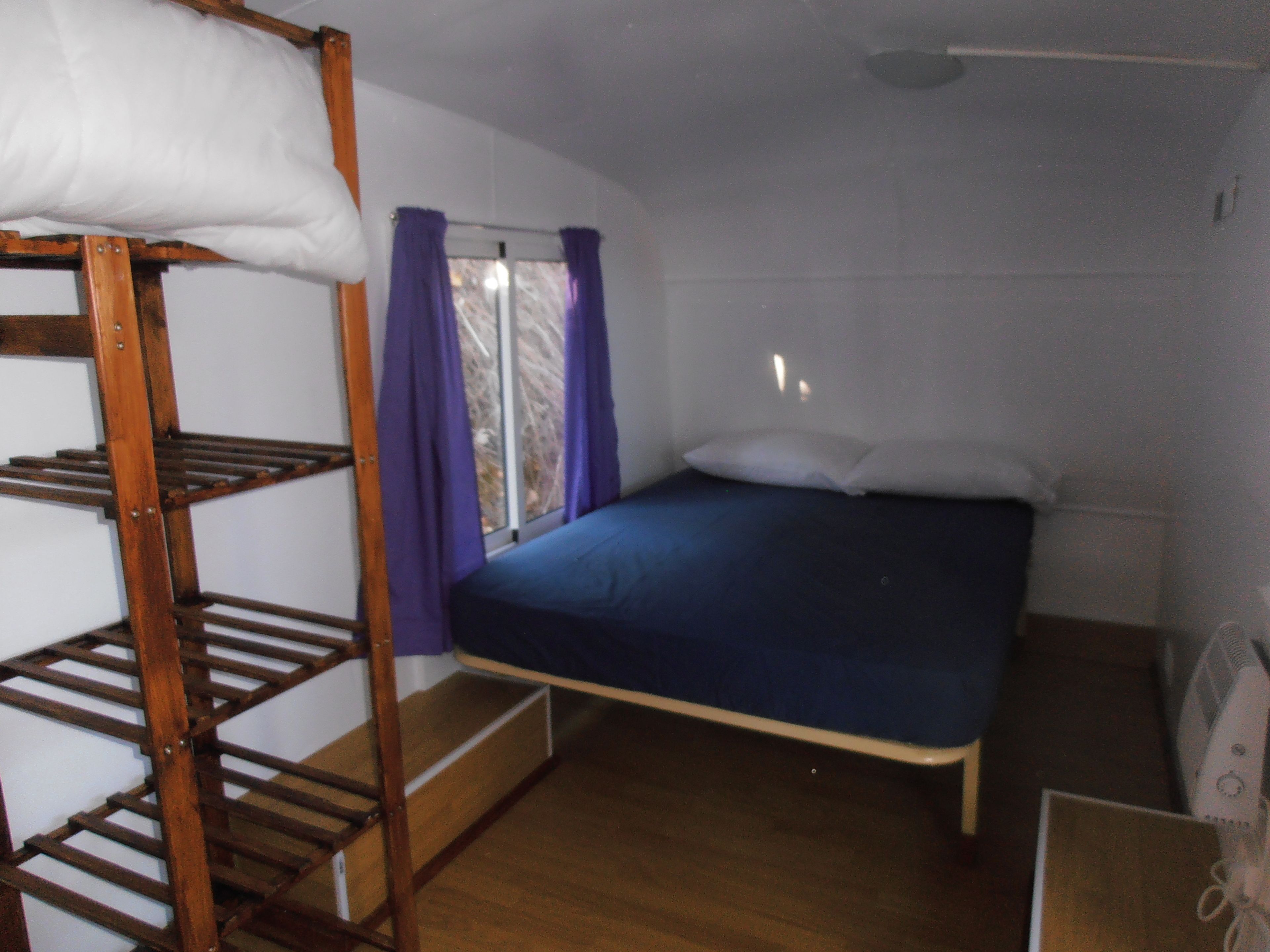 A bunk bed with a wooden ladder, a single bed with a blue mattress, purple curtains, and a window with a view of trees.