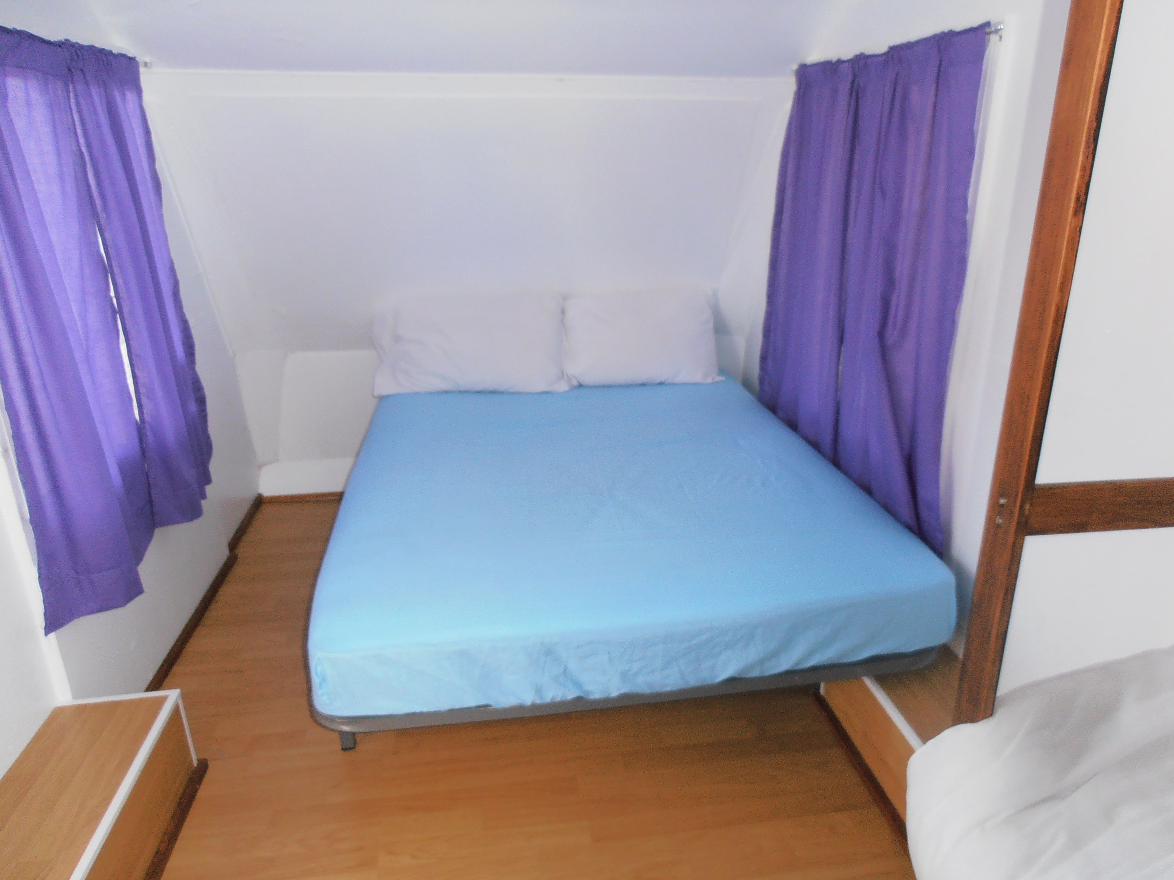 Una habitación pequeña con cama individual, colchón azul, almohadas blancas y cortinas púrpuras.