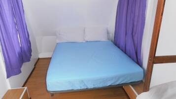 Una habitación pequeña con cama individual, colchón azul, almohadas blancas y cortinas púrpuras.