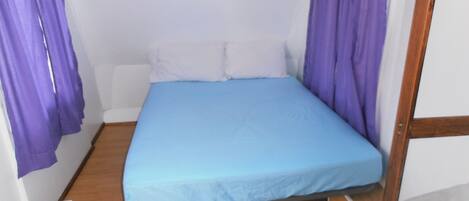 Una habitación pequeña con cama individual, colchón azul, almohadas blancas y cortinas púrpuras.