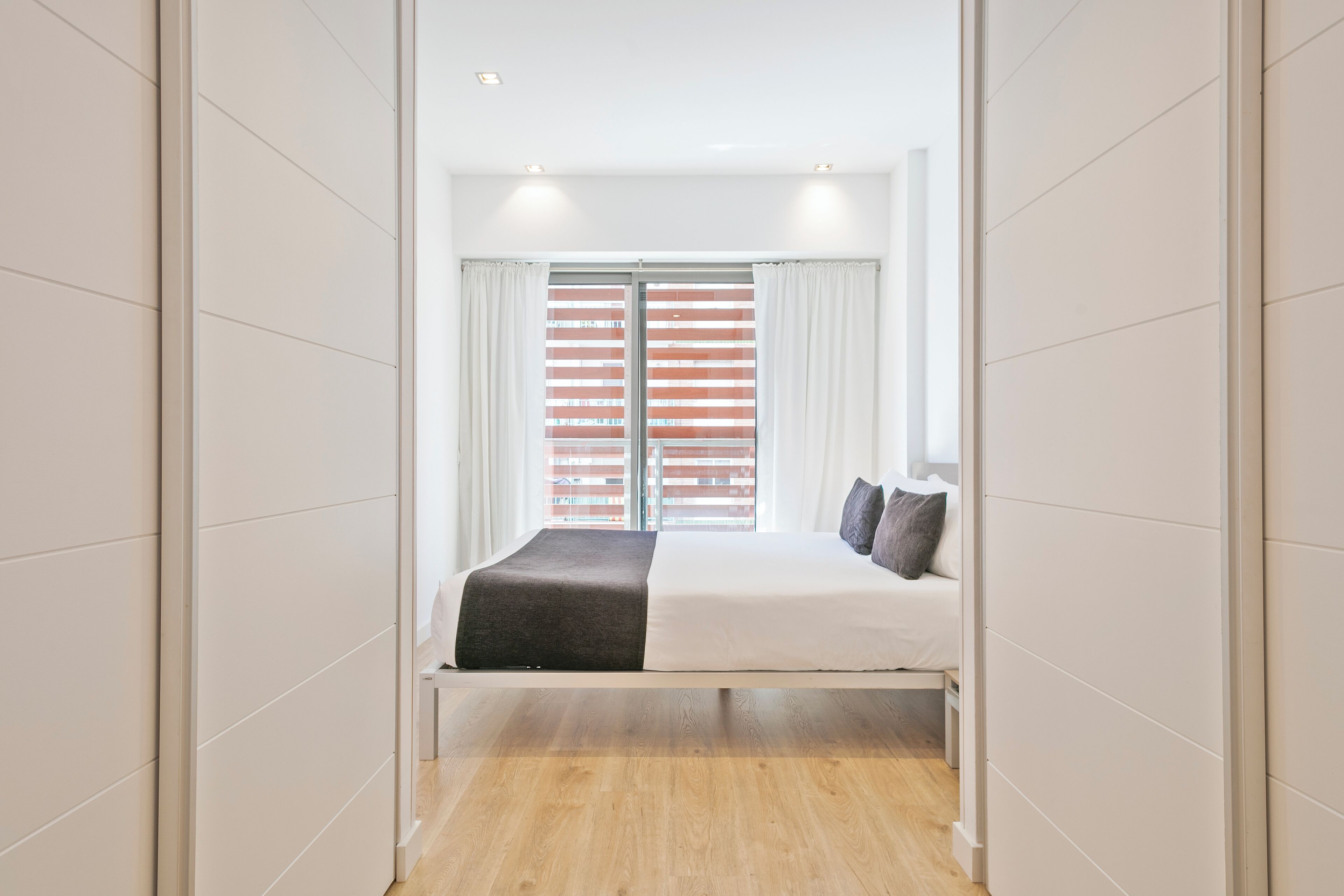 Een moderne slaapkamer met een bed, witte gordijnen en uitzicht door een raam met jaloezieën.