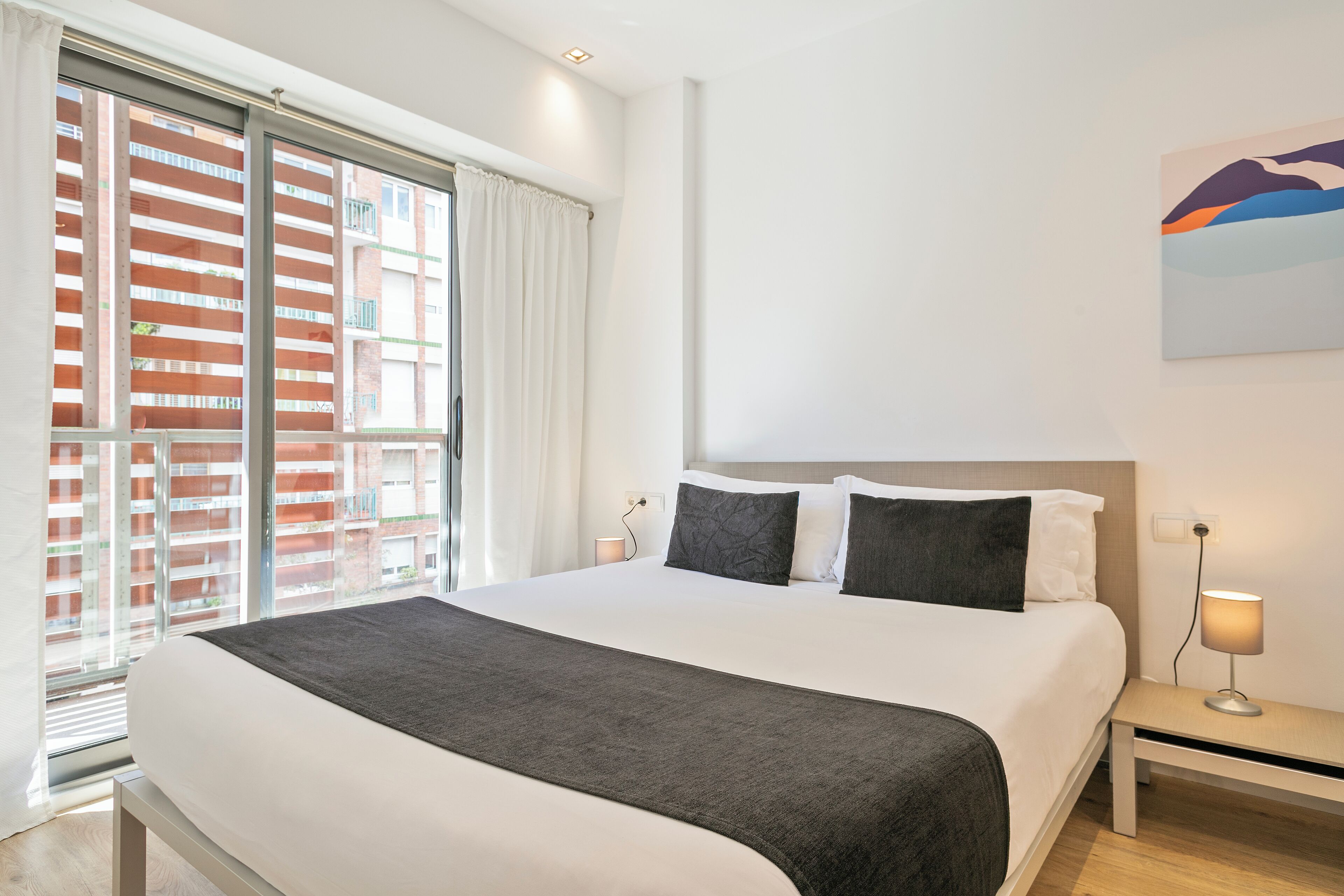 A modern hotel room with a large bed, a bedside table with a lamp, and a view of a cityscape through the window.