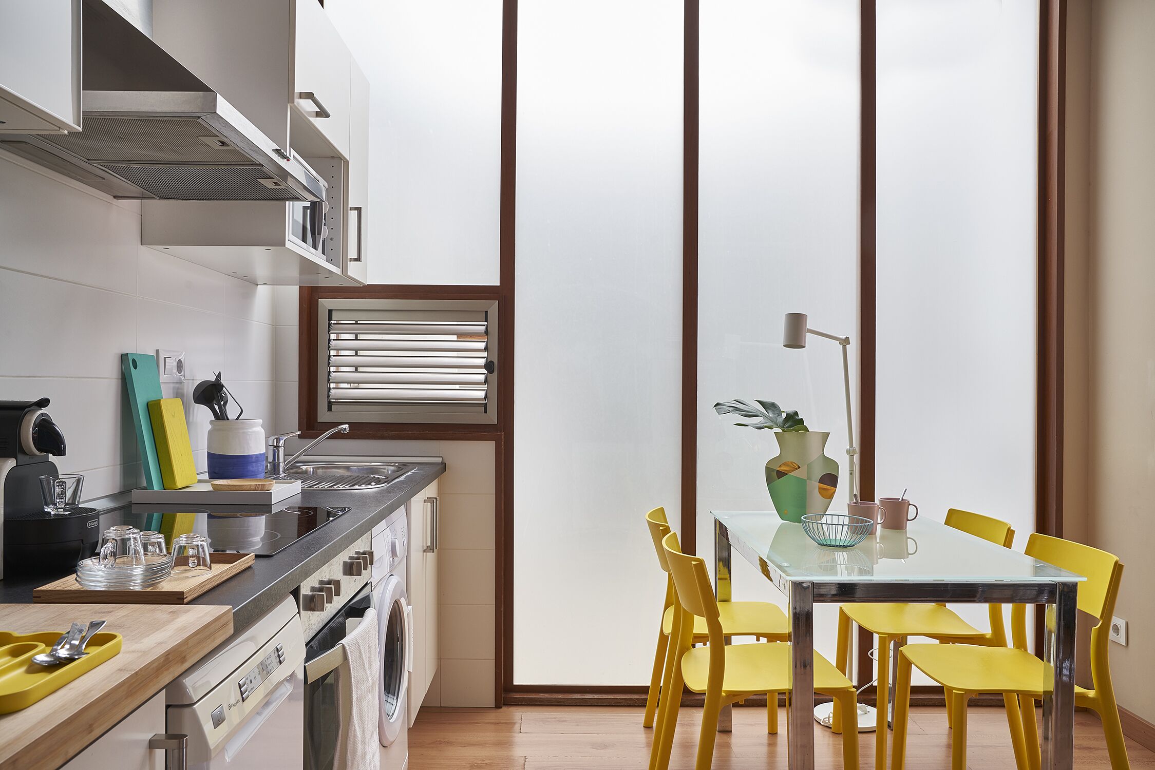 A compact kitchen with modern appliances, a dining area with yellow chairs, and a large window with blinds.