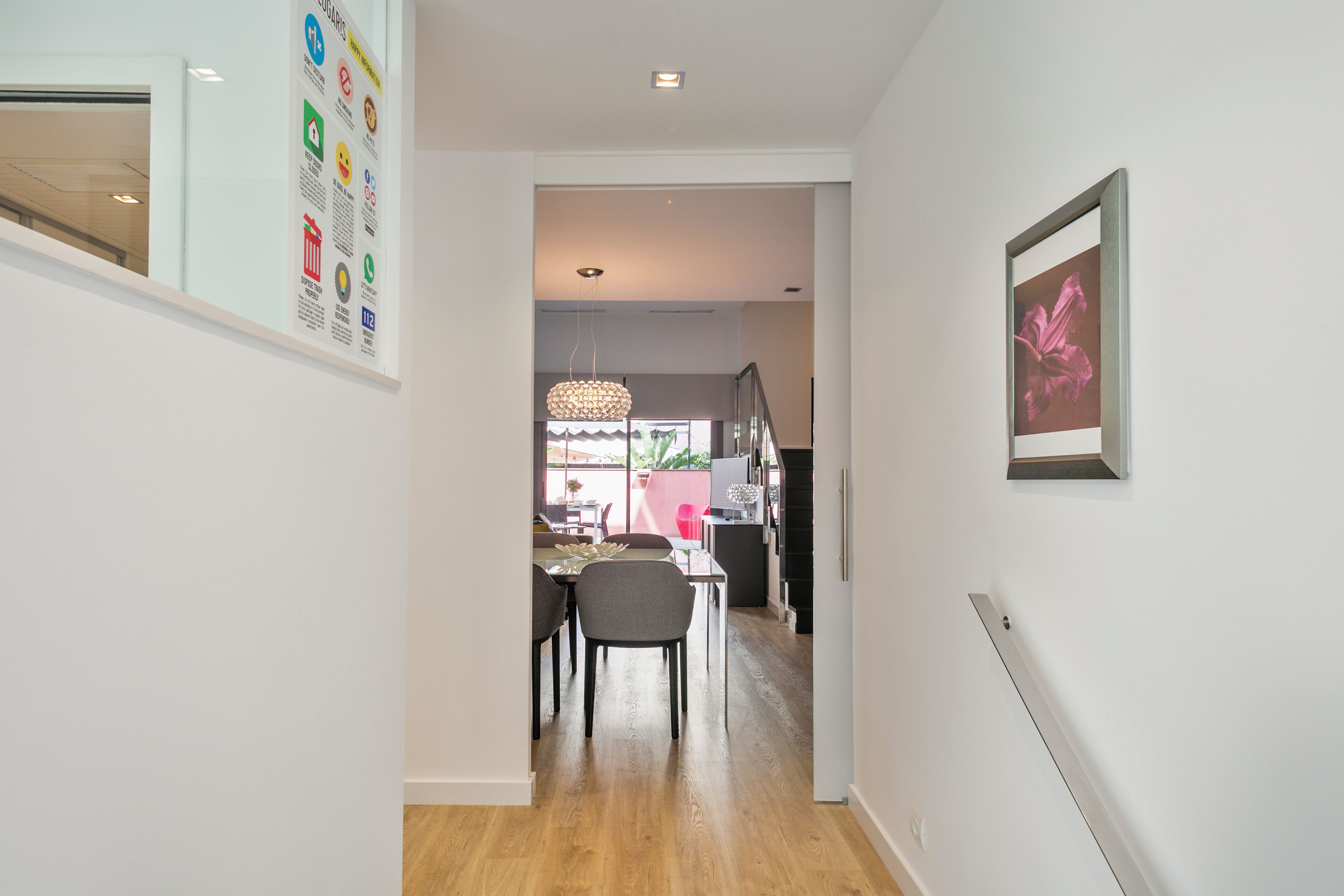 A hallway leading to a dining area with a table and chairs, a hanging light fixture, and a framed picture on the wall.