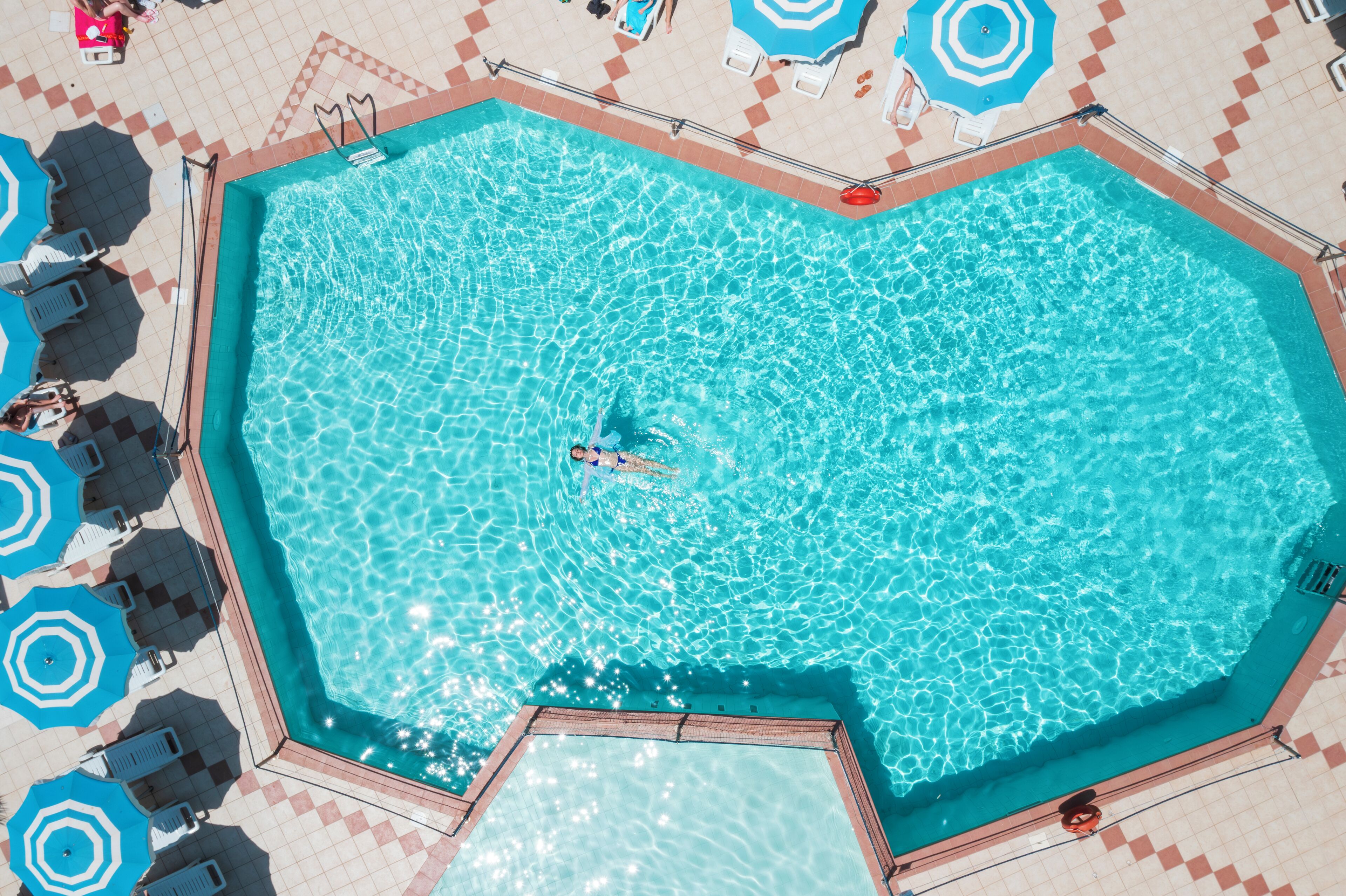 Una piscina grande y rectangular con agua azul cristalina, rodeada por una terraza con azulejos, sillas de descanso y sombrillas.