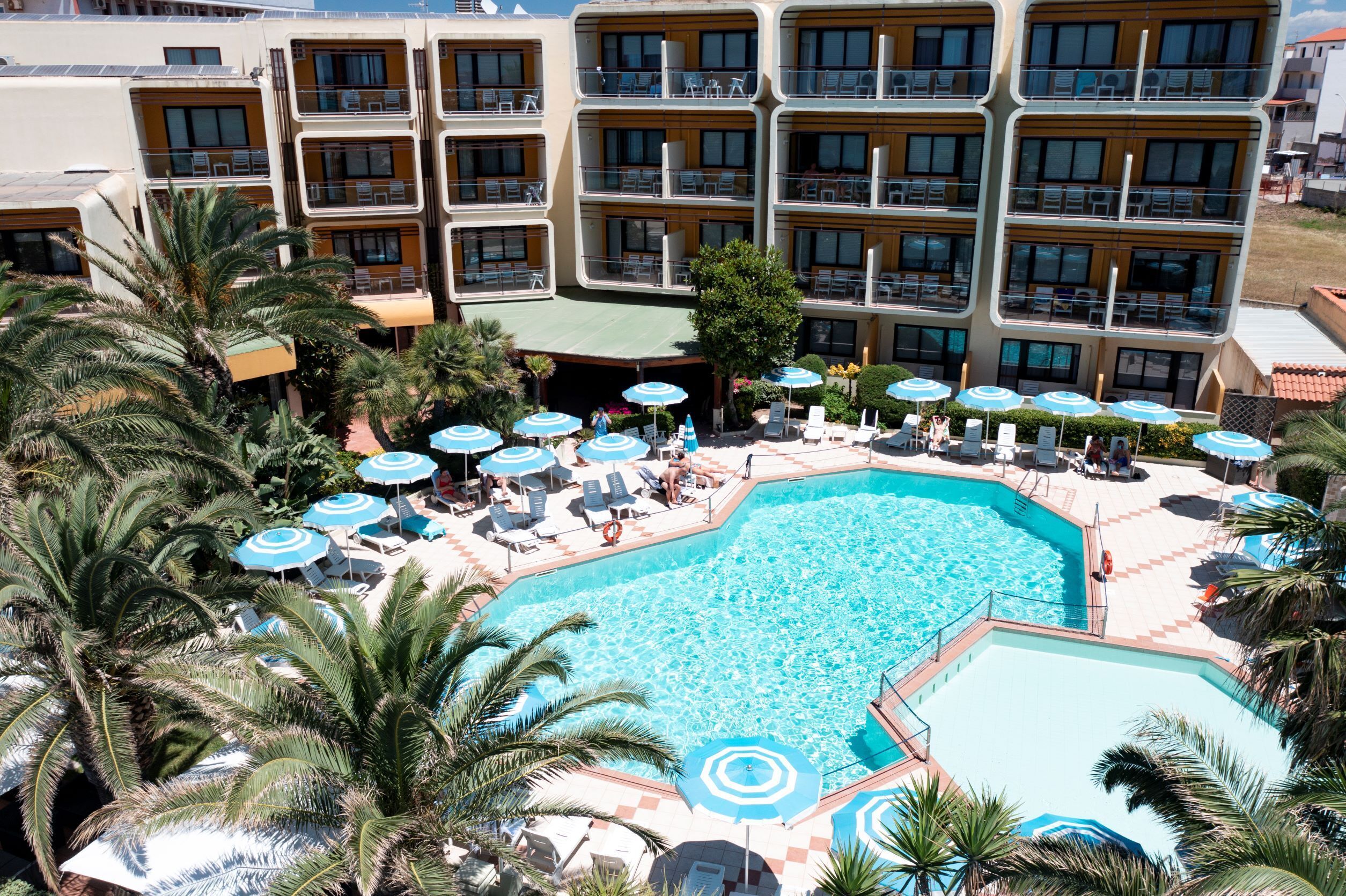 Un hotel con una amplia piscina rodeada de tumbonas y sombrillas, varios balcones y palmeras.