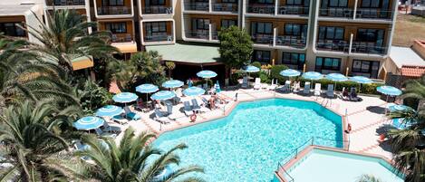 Un hotel con una amplia piscina rodeada de tumbonas y sombrillas, varios balcones y palmeras.