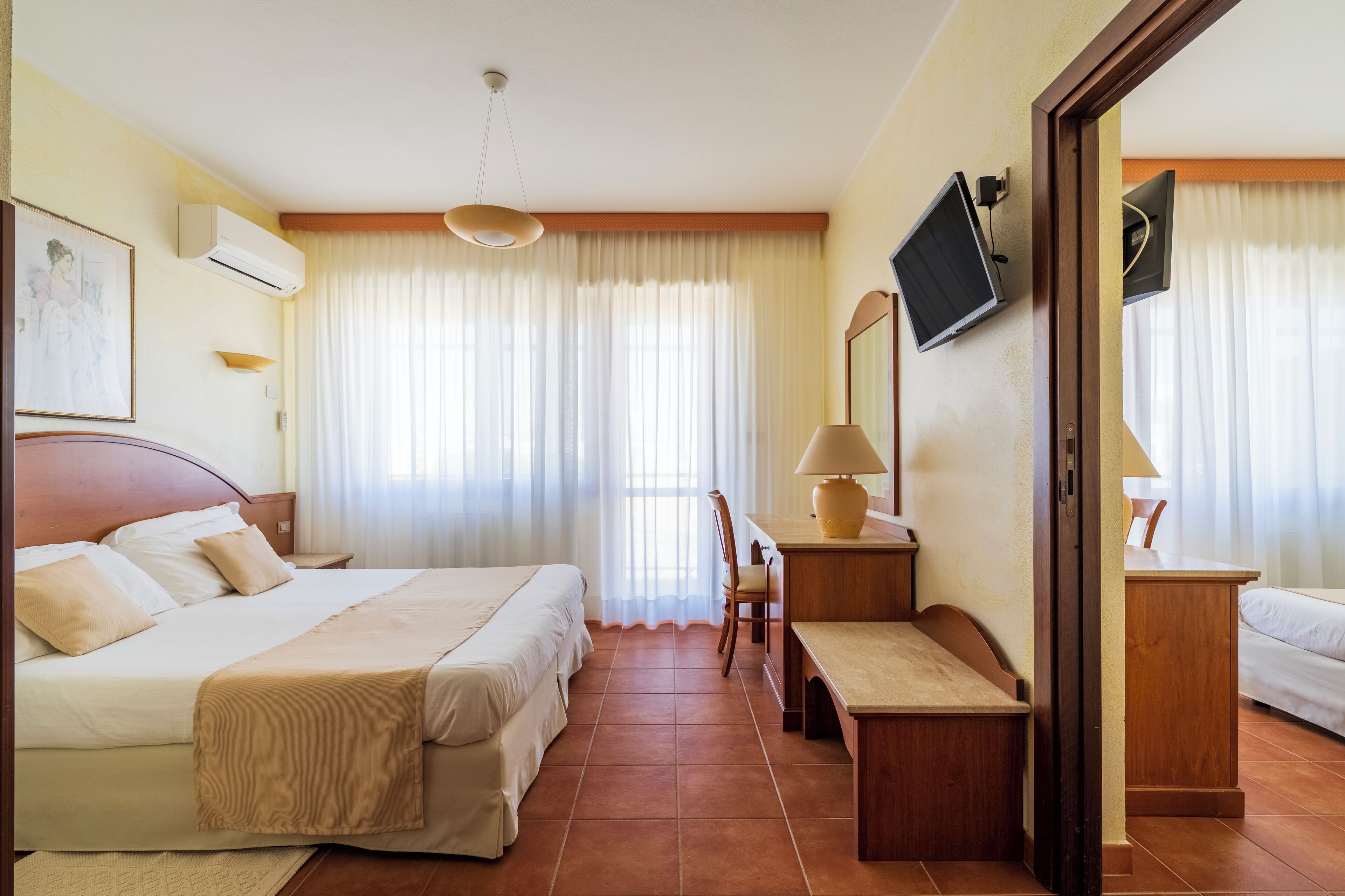 Una habitación de hotel con cama, mesitas de noche, escritorio, silla, televisor y ventana con cortinas.