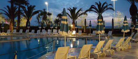 A poolside area with lounge chairs and umbrellas, surrounded by palm trees and a view of the sea.