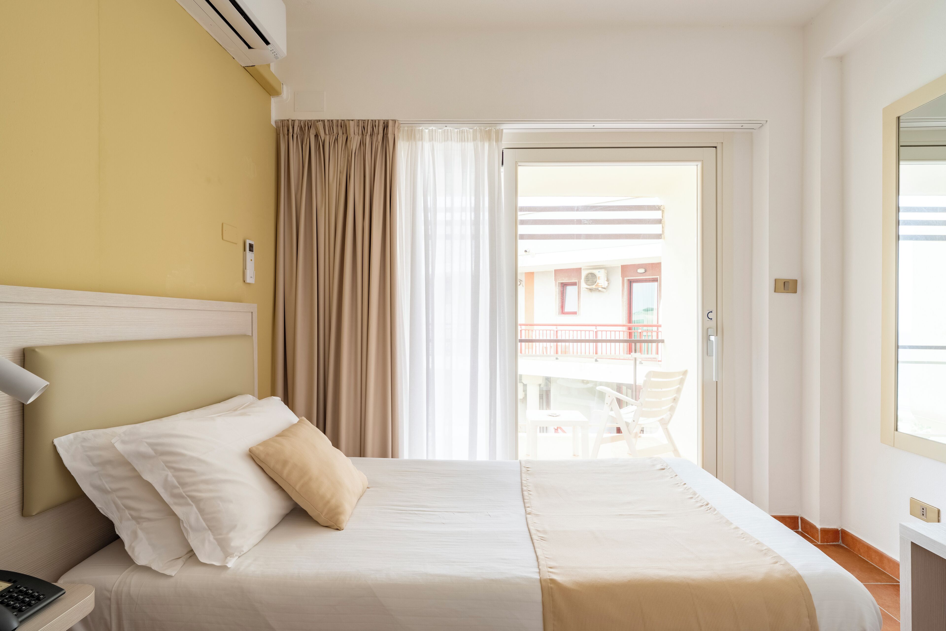 Una camera d'albergo con un letto, un comodino con una lampada, una finestra con tende e vista su un balcone esterno.