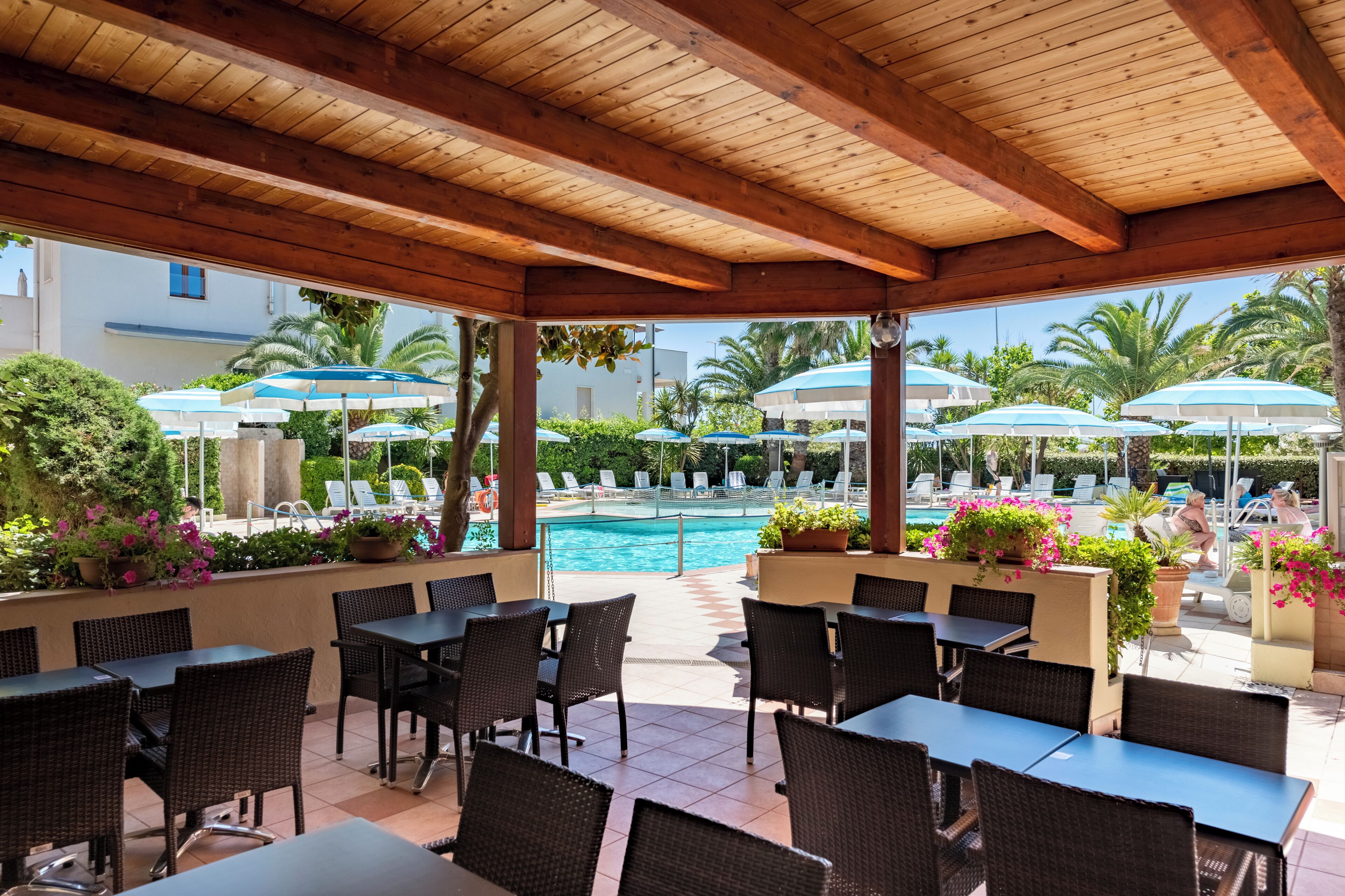 An outdoor patio area with wooden beams, tables, and chairs, overlooking a pool and lounge area with umbrellas.