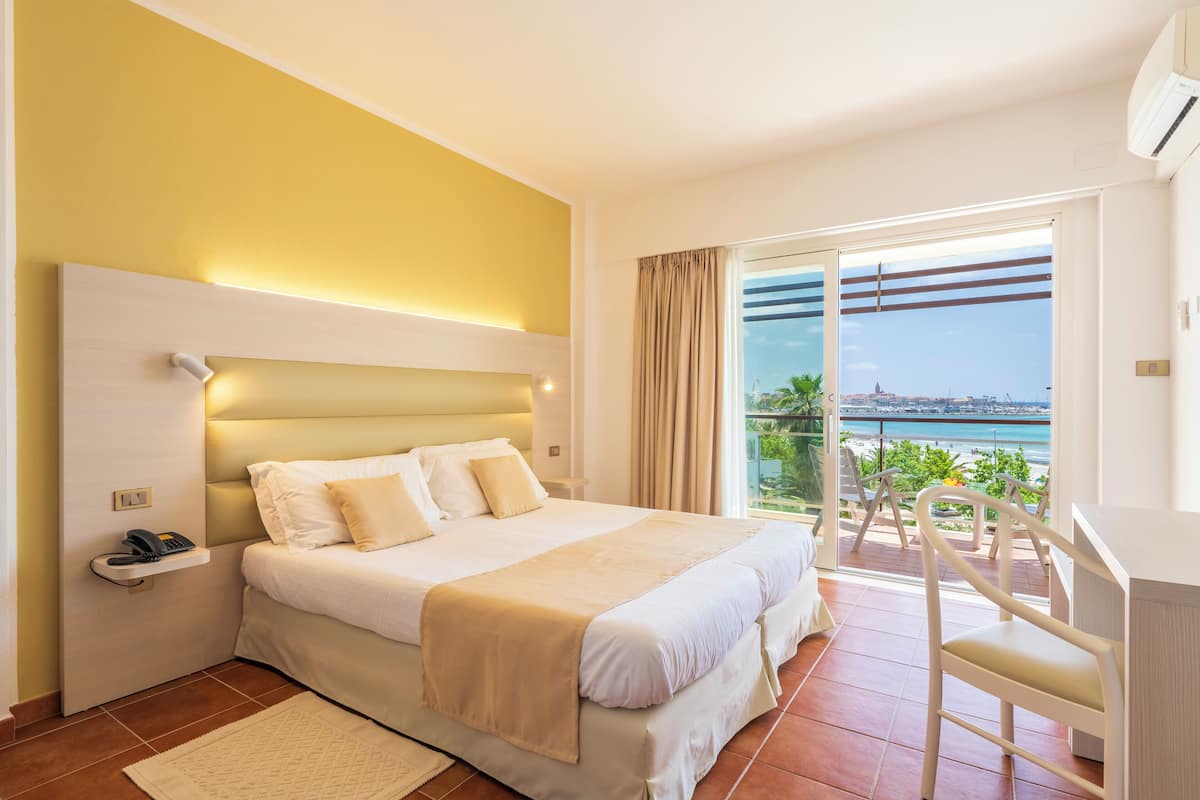 A hotel room with a large bed, a bedside table with a phone, a chair, and a view of the sea through a sliding door.