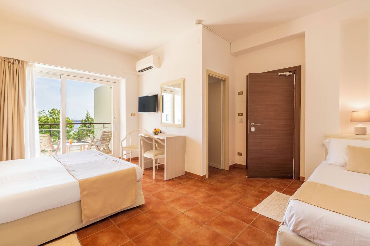 A hotel room with two beds, a desk, a TV, and a balcony with outdoor furniture.
