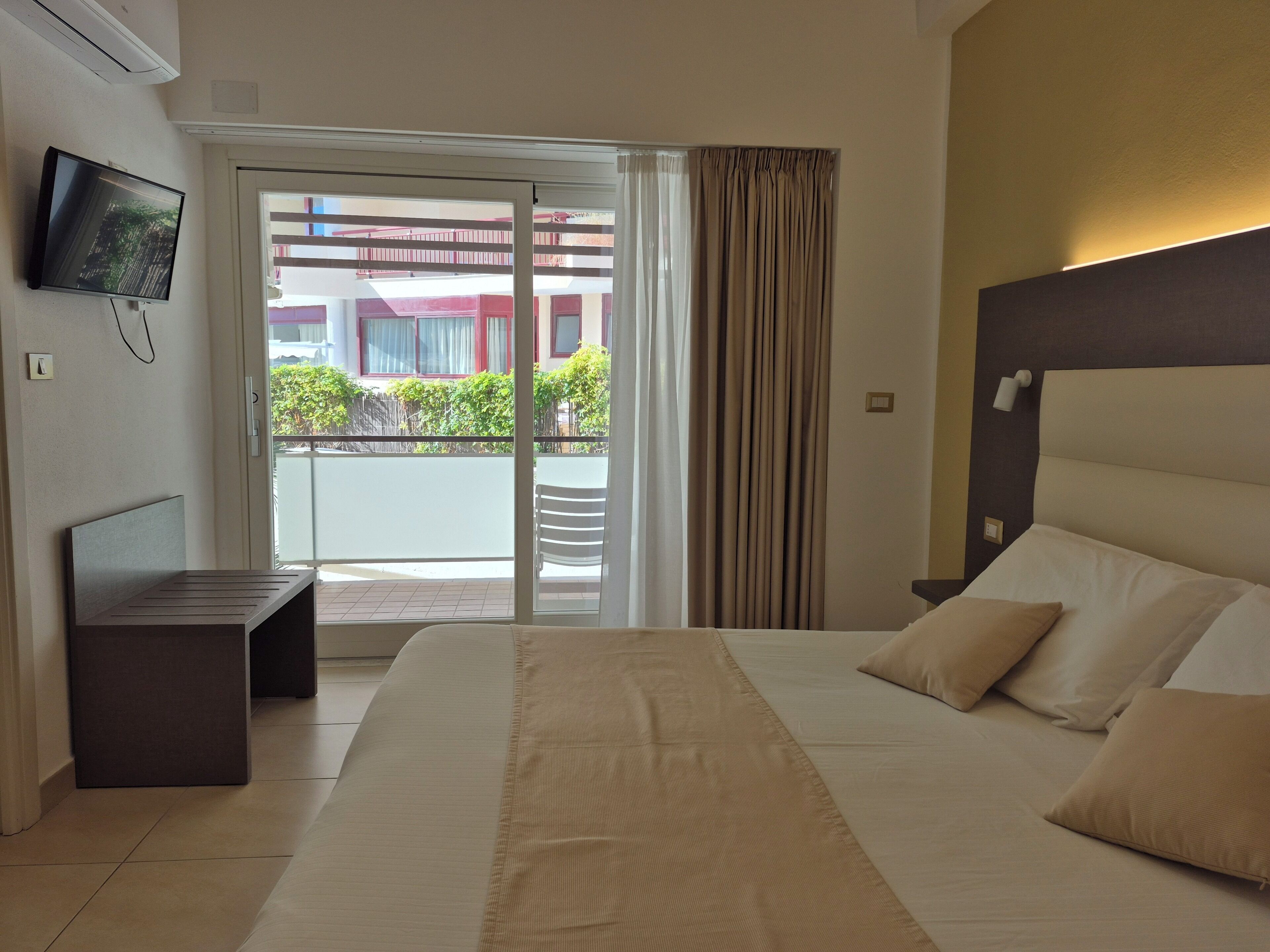 Quarto de hotel com cama, televisão presa na parede, porta de vidro deslizante para a varanda e cabeceira com luminária.