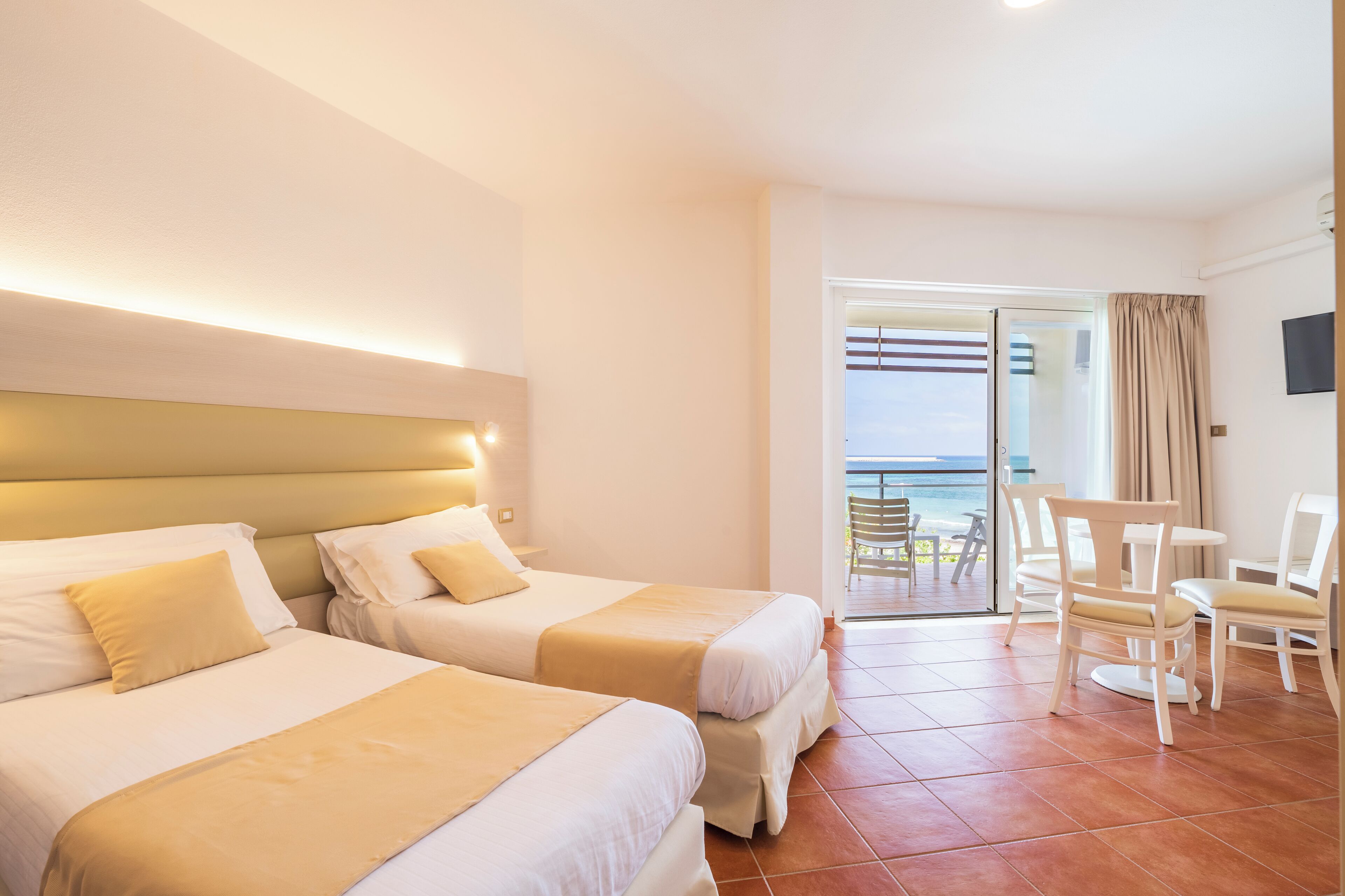 A hotel room with two beds, a balcony with a view of the sea, and a dining area with chairs.