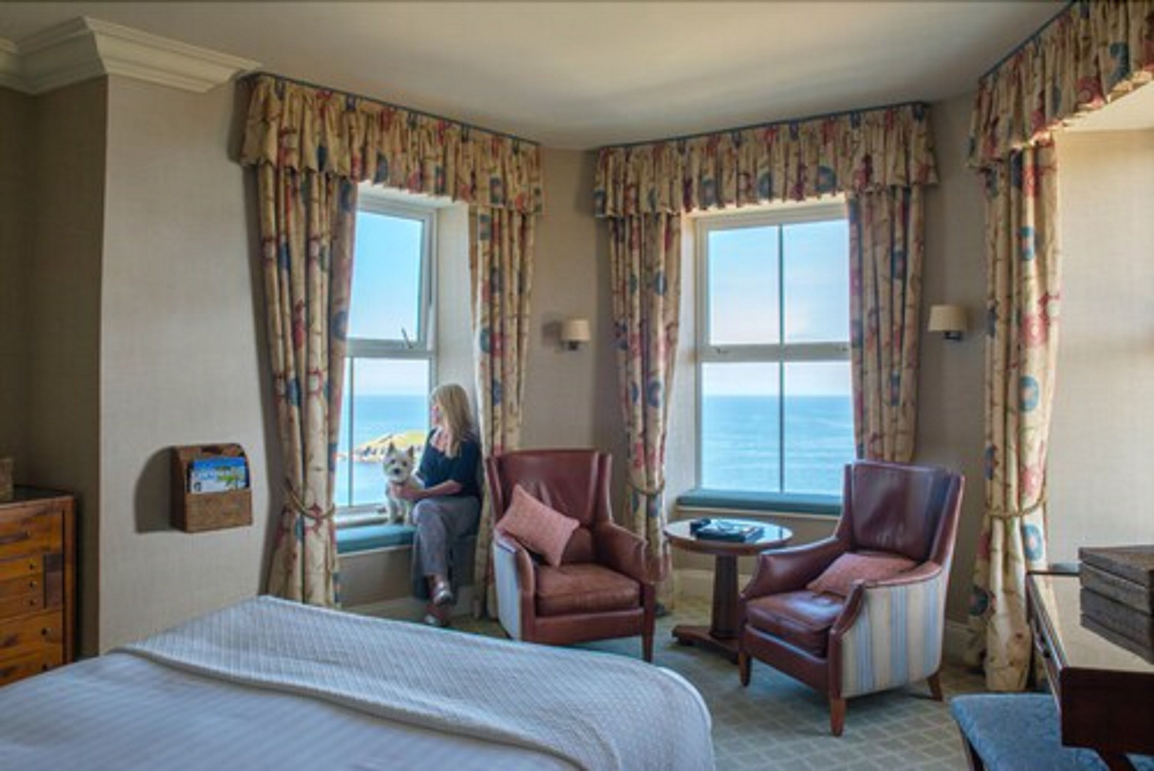 Habitación de hotel con una cama grande, dos sillones, una mesita y vistas al mar a través de dos ventanas.