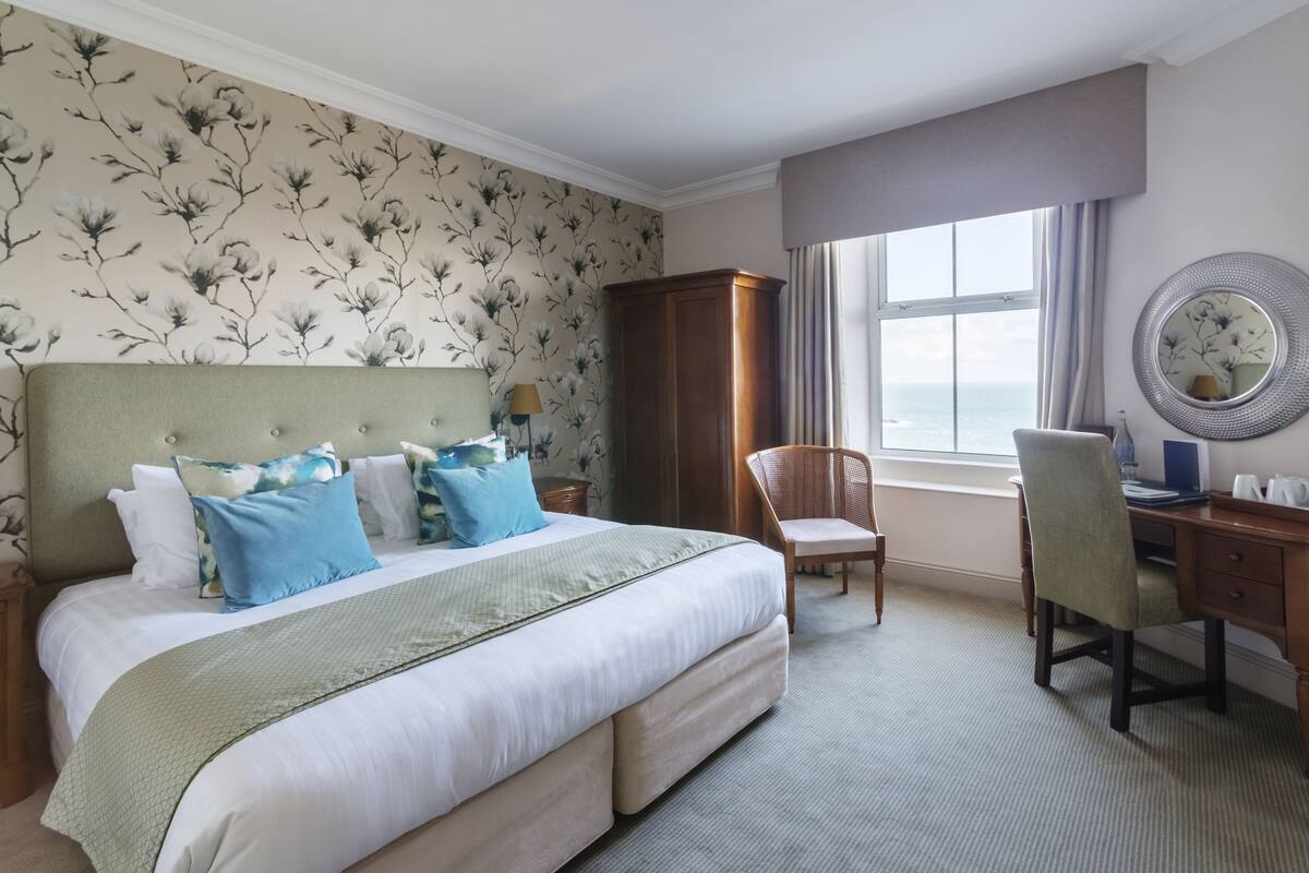 A hotel room with a large bed, a desk, a chair, a wardrobe, and a window with a view of the sea.