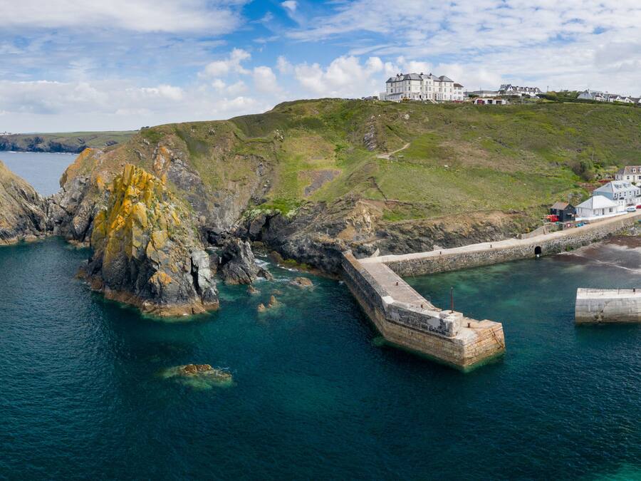 Uma vila costeira com um edifício branco imponente numa colina, um cais que se estende para o mar e uma costa rochosa.