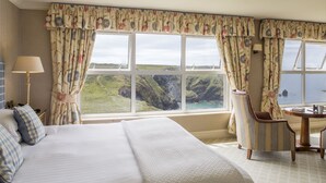A hotel room with a large bed, a chair, a table, and a view of a coastal landscape through the windows.