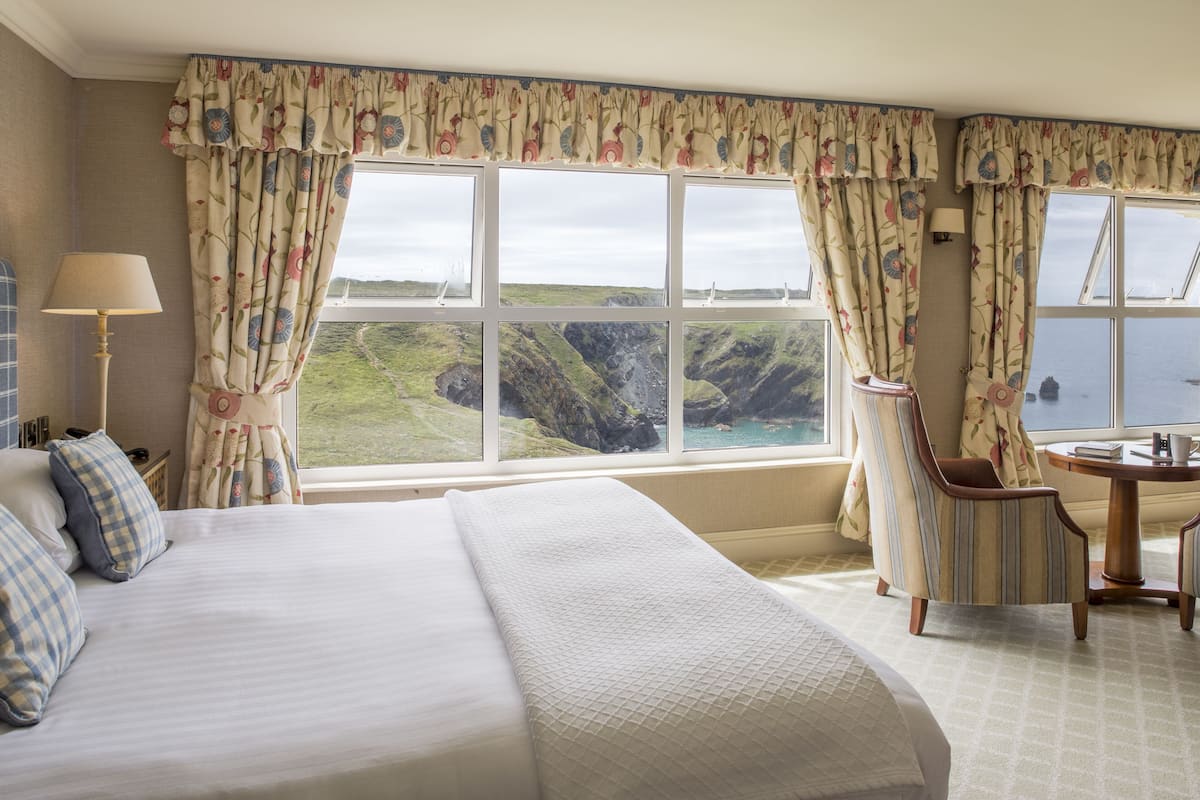 A hotel room with a large bed, a chair, a table, and a view of a coastal landscape through the windows.