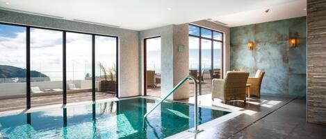 En moderne indendørs swimmingpool med glasvægge, havudsigt og et loungeområde med fletstole.