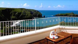 A balcony with a view of the ocean and cliffs, a wooden table with a bottle and glasses, and a white railing.