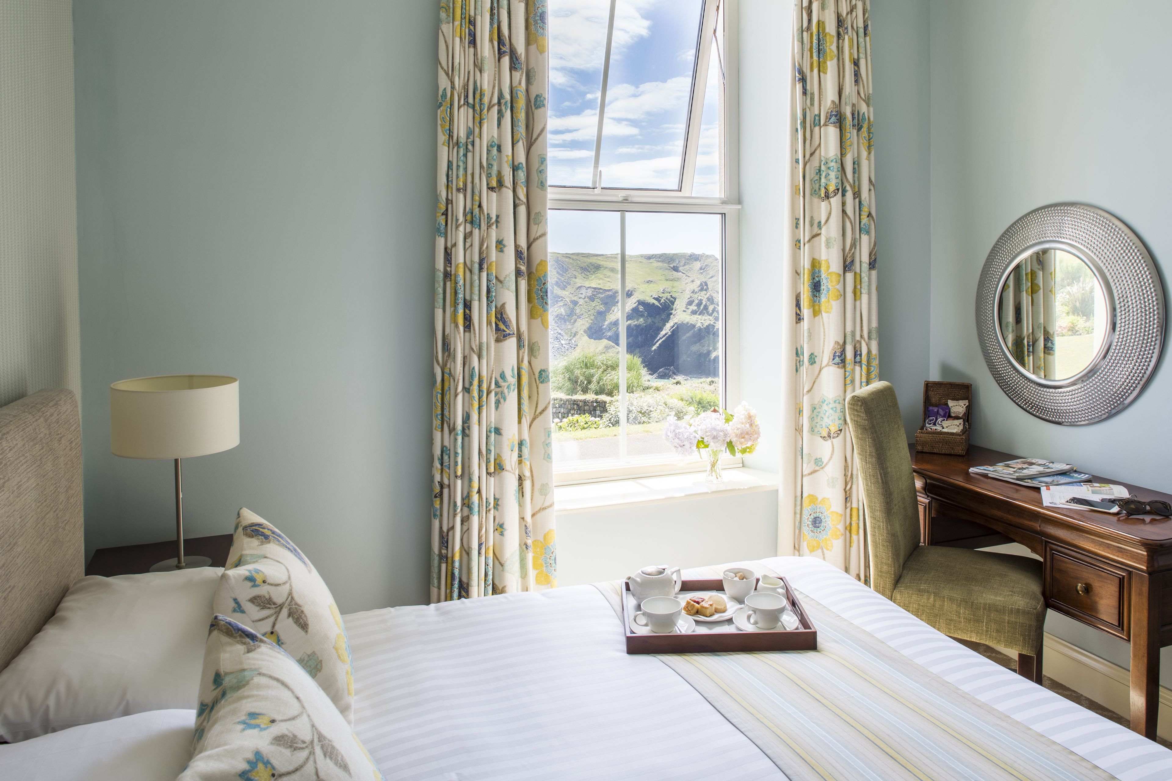 Una habitación de hotel con cama, escritorio, silla, ventana con cortinas y una bandeja con cosas para el desayuno.