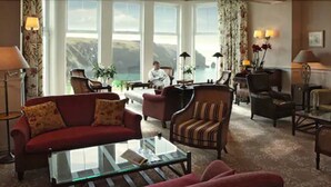 A spacious living room with a red sofa, a striped armchair, and a glass coffee table. There are multiple lamps and a view of the outdoors through large windows.