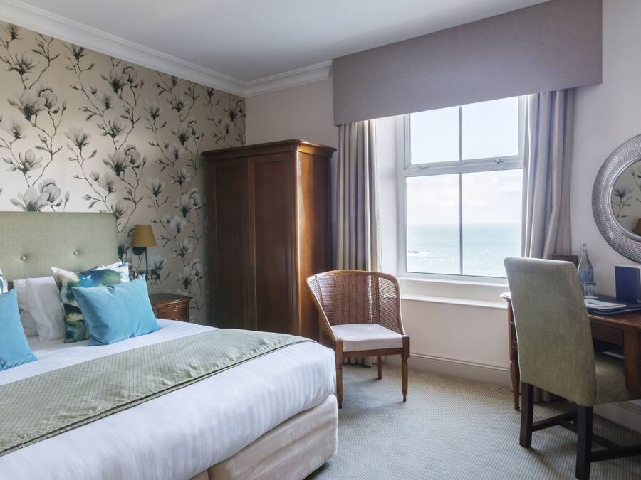 A hotel room with a bed, a desk, a chair, a wardrobe, and a view of the sea through a window.