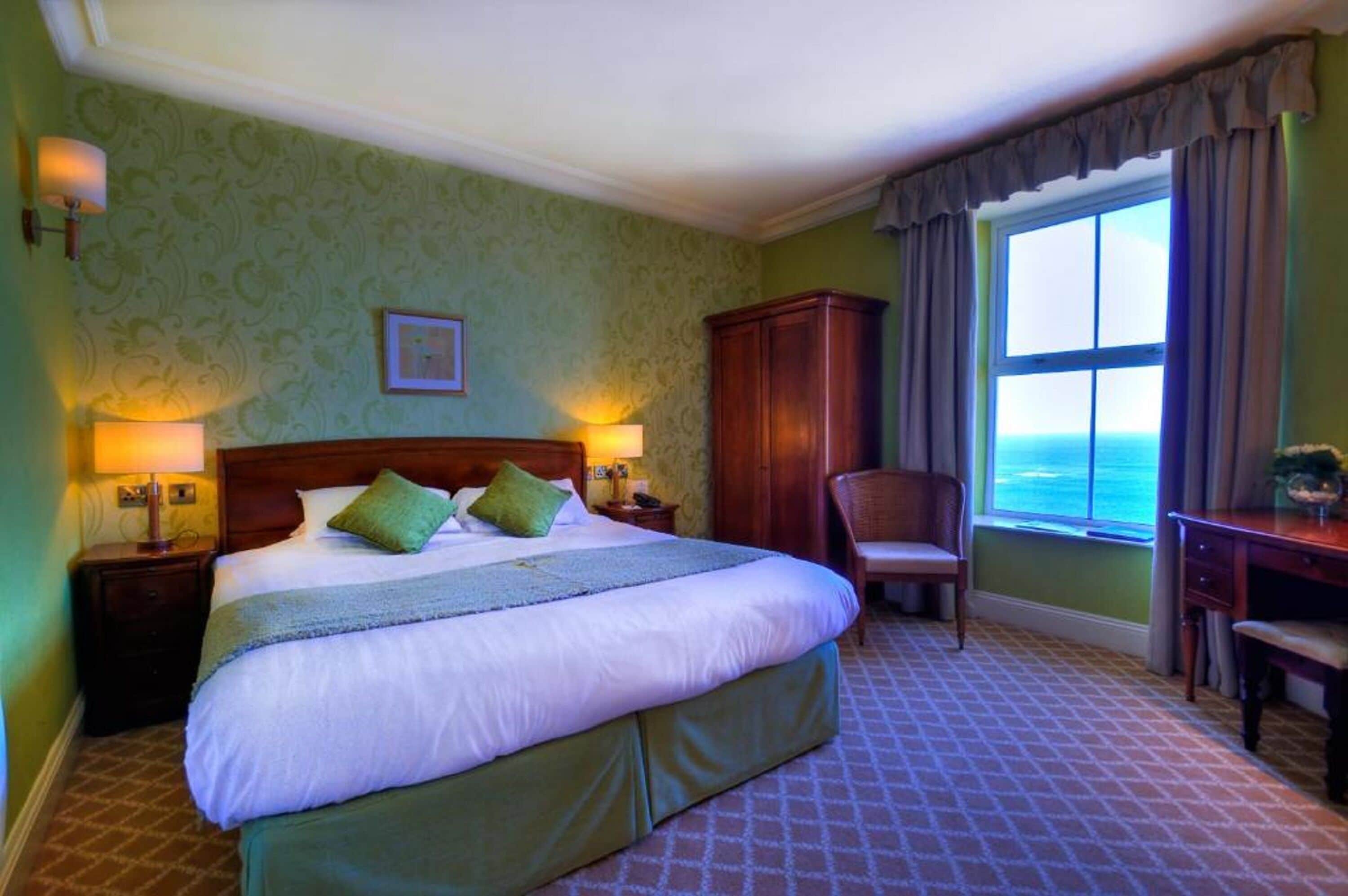 A hotel room with a large bed, two bedside lamps, a wooden wardrobe, a desk with a chair, and a window with a view of the sea.