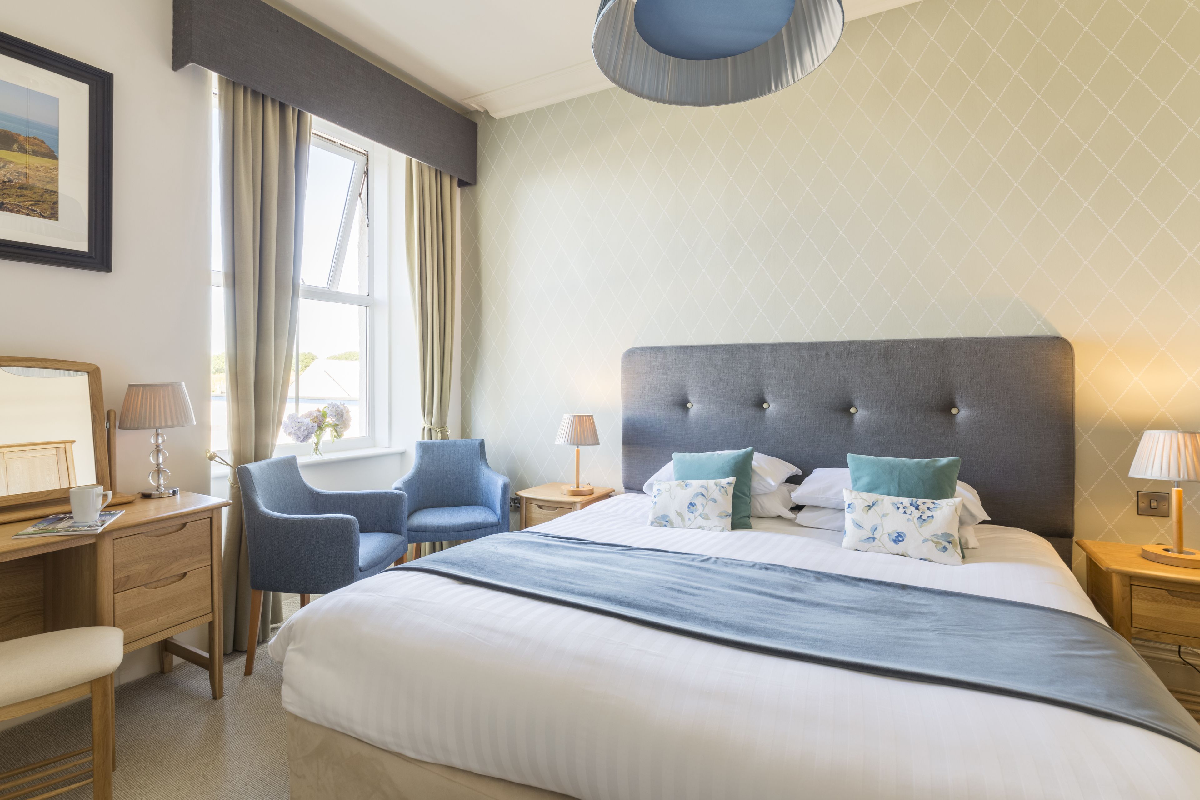A hotel room with a large bed, a desk with a chair, a window with curtains, and a bedside table with a lamp.