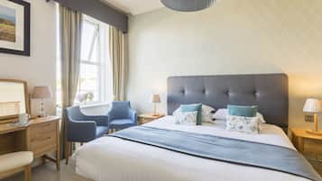 A hotel room with a large bed, a desk with a chair, a window with curtains, and a bedside table with a lamp.