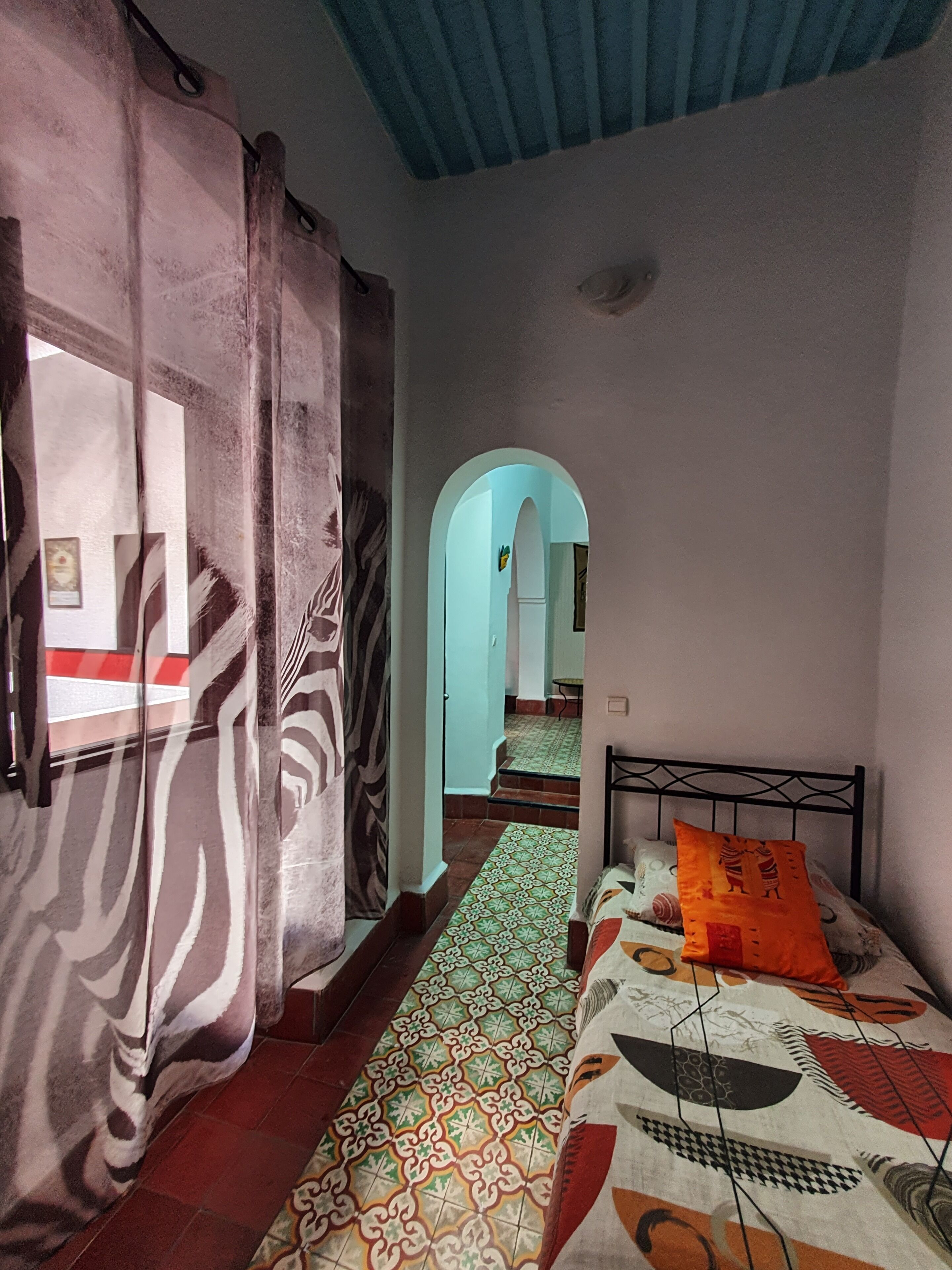 A room with a bed, a patterned rug, a window with curtains, and an archway leading to another room.