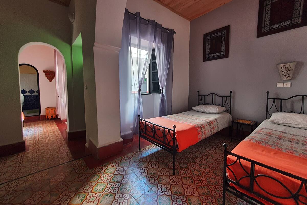 A room with two beds, a patterned floor, a wooden ceiling, and a window with curtains.