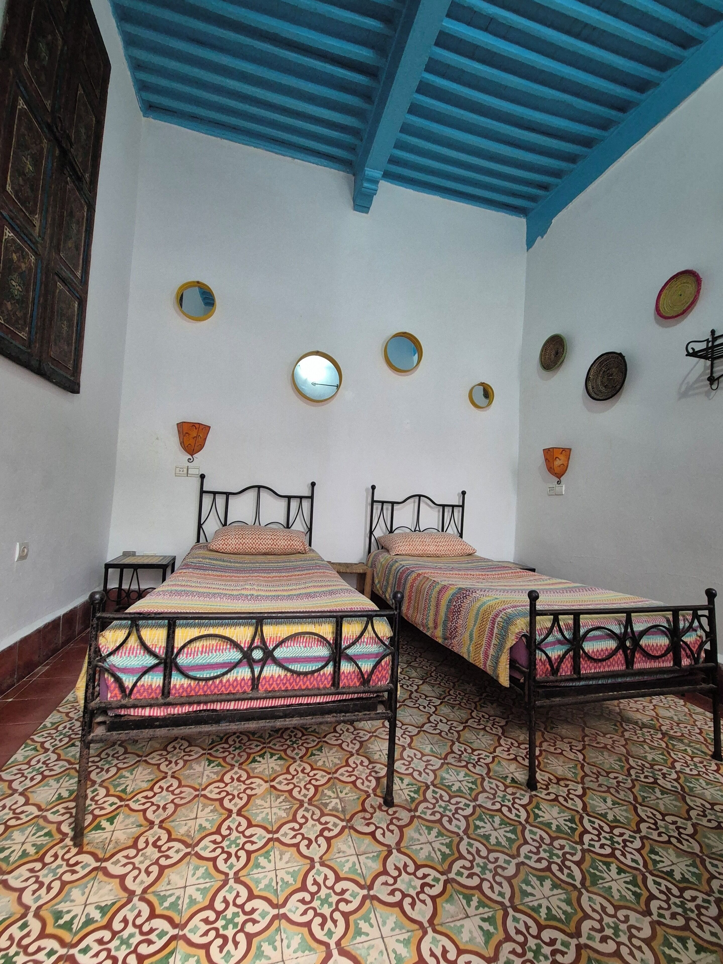 Habitación con dos camas, suelo con baldosas estampadas, techo azul y varias decoraciones en las paredes, incluyendo espejos y adornos.