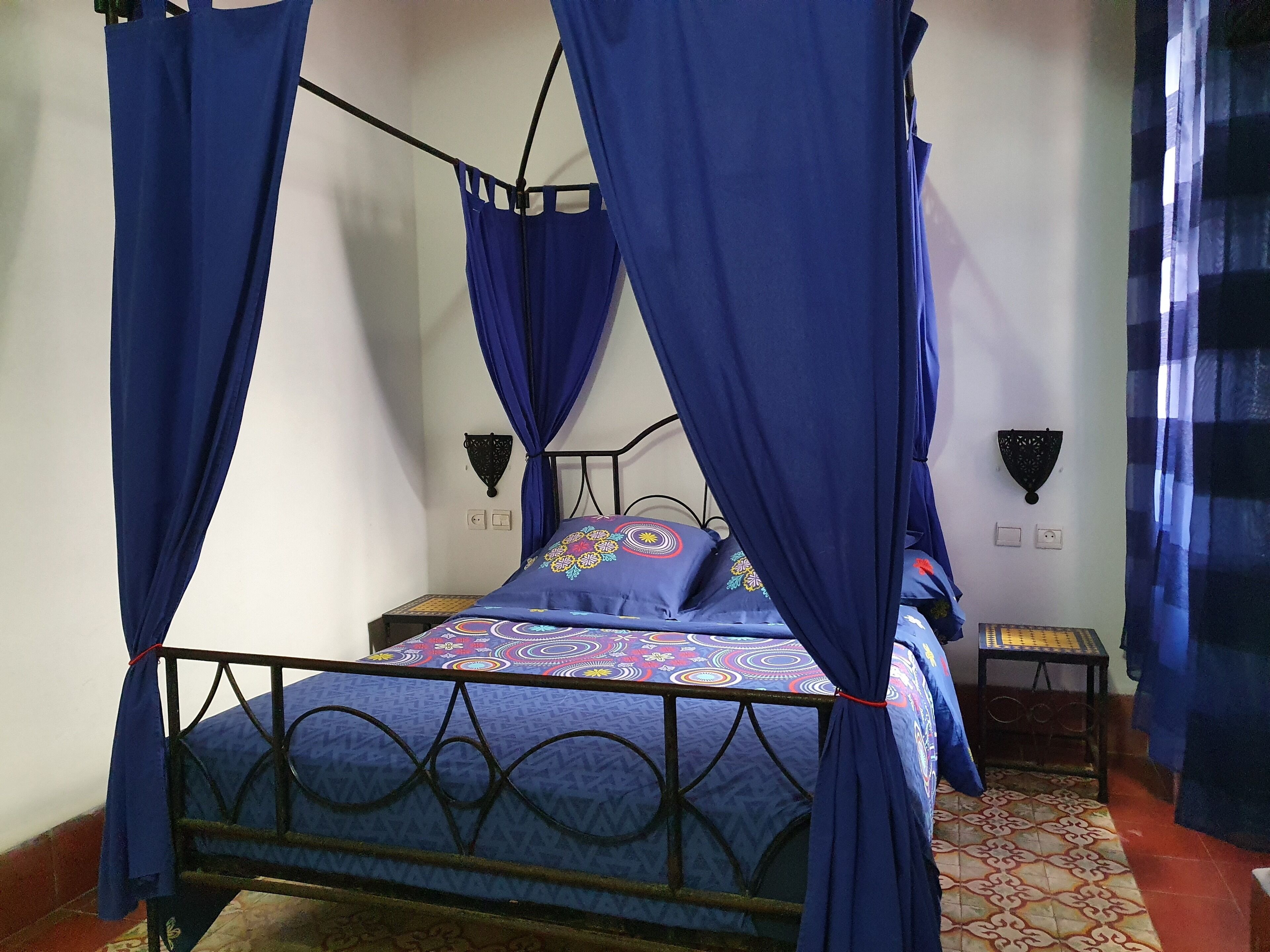 Una cama con estructura metálica y dosel azul, una colcha estampada, dos lámparas fijadas a la pared y una mesita de noche.