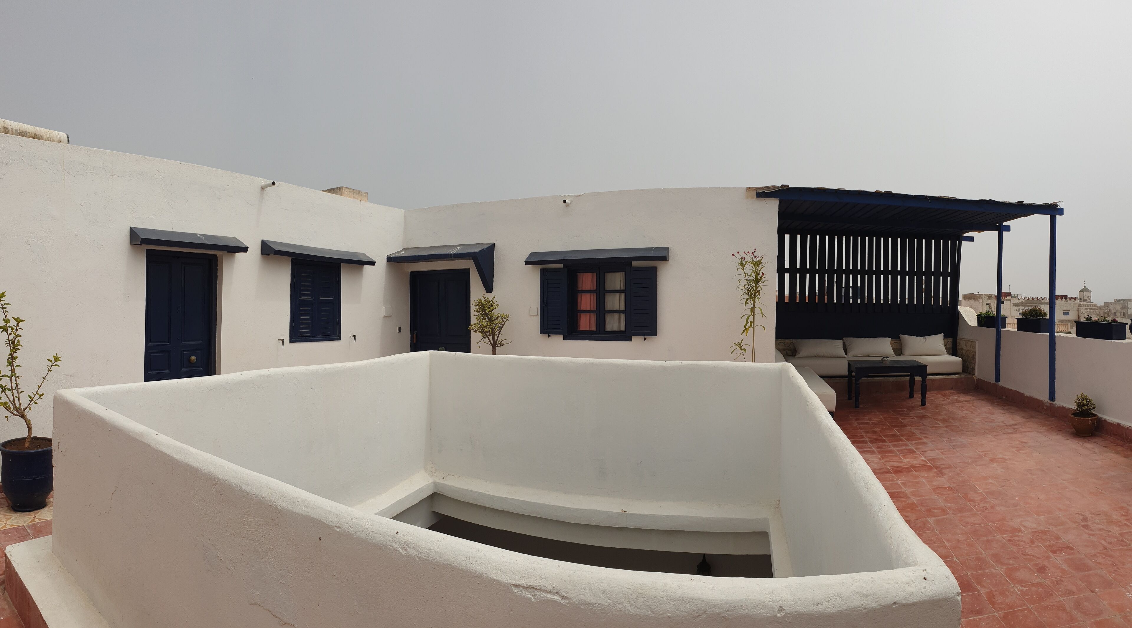 A rooftop terrace with white walls, dark shutters, and a seating area with a sofa and table.